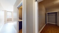 This single-floor space presents a thoughtful blend of modern finishes and industrial character throughout its layout. The kitchen serves as a standout feature, offering white cabinetry with dark hardware, dark granite countertops, a kitchen island, white subway tile backsplash, stainless steel appliances, and open wooden shelving suspended by black metal rods with pendant cage lighting overhead. Natural light pours into the adjoining living and dining area through large windows framed in dark trim, which also reveal views of the surrounding urban streetscape. A partial exposed brick wall adds warmth and texture to this bright, open area, complemented by light hardwood-style flooring that runs throughout the space.

The hallway connects the living areas to the private quarters, featuring a sliding barn door mechanism and clean white trim work. The bedroom is generously sized, finished with warm hardwood-style flooring and neutral gray walls, and includes two separate reach-in closets with built-in shelving and hanging rods — offering ample storage. The bathroom is well-appointed with a dark-framed glass-enclosed shower, a dark vanity, and matching hardwood-style flooring, maintaining a cohesive design aesthetic. Overall, the space balances contemporary style with practical functionality.