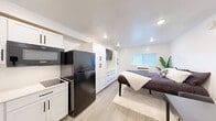 This efficiently designed studio unit offers a thoughtfully organized open-plan layout that maximizes every square foot. The interior features light wood-look flooring throughout, crisp white walls, and recessed ceiling lighting that creates a bright, clean ambiance across the entire space.

The kitchen is well-appointed with white shaker-style cabinetry accented by matte black hardware, a stainless steel sink with a black pull-down faucet, a smooth-top electric cooktop, an over-the-range Whirlpool microwave, and a full-size black refrigerator. Ample upper and lower cabinet storage ensures excellent functionality for daily use.

A wall of built-in white cabinetry with multiple drawers and upper cabinets serves as a central organizational feature of the main living area, with a wall-mounted television integrated seamlessly into the unit. The sleeping area accommodates a full-size bed and benefits from natural light provided by a window fitted with horizontal blinds and a wall-mounted air conditioning unit beneath it.

A compact dining area with a dark-toned table and two chairs is positioned near the sleeping space, making efficient use of the open floor plan. A full-length mirror near the bathroom entry adds depth to the space. The bathroom includes a vanity with a white basin sink and overhead lighting. The overall design is modern, minimalist, and move-in ready.