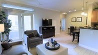 This single-floor residence offers an open-concept layout that seamlessly connects the living, dining, and kitchen areas, creating a cohesive and functional living environment. The living area is carpeted and features recessed lighting complemented by crown molding, lending a polished, finished look to the space. A glass-paneled door floods the room with natural light and provides direct access to an outdoor balcony, enhancing the sense of openness.

The living area accommodates a comfortable seating arrangement centered around a round mirrored coffee table and an upholstered ottoman, with a media console providing practical storage. Neutral wall tones throughout create a calm, welcoming atmosphere that pairs well with the light-colored carpet underfoot.

Adjacent to the living area, a dining space features bar-height seating, making it ideal for casual meals or entertaining. A hallway visible from the main living area suggests additional rooms beyond, including what appears to be a bathroom. Recessed and overhead lighting throughout ensures the space remains well-lit, while the overall layout promotes easy flow and versatility.