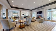 This single-floor community amenity space is thoughtfully designed to offer a variety of functional and social environments. The central lounge area serves as the heart of the space, featuring a warm fireplace framed by a wall-mounted television, stylish patterned armchairs and sofas arranged around wooden coffee tables, and a striped area rug that anchors the seating. Large windows dressed with sheer curtains flood the room with natural light, while recessed lighting and a bold dark tray ceiling add architectural character. A set of glass doors opens directly to an outdoor patio area, enhancing the indoor-outdoor connection. Adjacent to the lounge, a modern kitchen showcases dark cabinetry with sleek hardware, white quartz countertops, a classic subway tile backsplash, hardwood-style flooring, and multiple windows overlooking greenery. A wall-mounted TV and ample counter space make it ideal for gatherings. The business center offers dual workstations with computers, complemented by warm wall sconces and wood-look flooring. A private conference room features a solid wood table, textured wall panels, and curated abstract artwork. The game and social lounge boasts soaring ceilings, expansive windows, bar-height tables, and a shuffleboard table, creating an energetic yet welcoming atmosphere throughout.