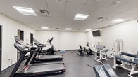 Located at 136 N Third St in Hamilton, Ohio, this fitness facility offers a generously sized, single-floor workout space designed to accommodate a wide variety of exercise needs. The interior features a clean, functional aesthetic characterized by white walls, a drop-panel ceiling with recessed fluorescent lighting panels, and durable dark rubber flooring throughout — a practical choice for high-traffic fitness environments.

The open layout is organized to maximize usability, with clearly defined zones for cardio and strength training. The cardio area is equipped with multiple Sole-brand treadmills, an elliptical trainer, and a recumbent stationary bike, providing options for users at varying fitness levels. The strength training section features an extensive array of equipment, including cable machines, selectorized weight stations, benches, a squat rack with barbell storage, and free weights, offering a thorough range of resistance training options.

Wall-mounted flat-screen televisions are positioned throughout the space to enhance the workout experience. The room benefits from multiple entry points and is well-ventilated with visible HVAC units in the ceiling. The overall ambiance is bright, clean, and organized, conveying a professional fitness environment suited for comprehensive training routines.