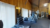This fitness center combines a rustic, inviting aesthetic with a comprehensive selection of exercise equipment. The interior is defined by rich, warm wood paneling that lines the walls and extends across the vaulted ceiling, creating an enveloping, cabin-like atmosphere that sets it apart from a typical gym environment. The angled ceiling adds a sense of spaciousness and architectural character to the single-floor facility. The workout area is well-appointed with a full dumbbell rack offering a wide range of free weights, a rowing machine, an elliptical trainer, and a multi-station strength machine, providing users with options for both cardiovascular and resistance training. A weight bench is conveniently positioned nearby for targeted exercises. Large mirrors mounted along one wall enhance the sense of space and allow users to monitor their form during workouts. The flooring features durable, cushioned gym matting suited for high-impact activity. Vertical blinds on the windows provide control over natural light, allowing the ambiance to be adjusted throughout the day. Overall, the space delivers a functional and comfortable fitness environment with a distinctly warm and natural character.