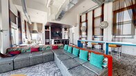 Located in the heart of downtown St. Louis at 219 N Eighth St, this generously sized amenity space offers a dynamic blend of industrial-chic architecture and vibrant contemporary design. The main lounge area impresses with soaring ceilings, exposed ductwork, and floor-to-ceiling windows that frame sweeping views of the city skyline and the iconic Gateway Arch. Bold furnishings — including modular gray sectional sofas accented with teal and red throw pillows — are complemented by patterned carpeting and a bold turquoise bar counter with wood-topped stools. A living moss wall installation adds a striking natural focal point near a bright, well-appointed secondary lounge with colorful seating and city views. The game room features exposed brick walls, a pool table, and café-style bistro tables, retaining the raw character of the building's architectural heritage. The kitchen and bar area offers dark cabinetry, stainless steel appliances, and bar seating. Outside, an expansive rooftop terrace delivers panoramic downtown views, complete with a spa pool, colorful lounge chairs, covered pergola seating, stainless steel cable railings, and lush planters — providing an ideal outdoor gathering environment above the St. Louis skyline.