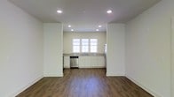 This single-floor space showcases a clean, modern design with an open-concept layout that seamlessly connects the living area to the kitchen. The flooring throughout is rich, dark hardwood, providing a warm contrast to the bright white walls and trim.

The kitchen is positioned at the far end of the open space and is outfitted with white shaker-style cabinetry, light-toned countertops, and stainless steel appliances, including a dishwasher and refrigerator. A trio of windows above the sink fills the kitchen with natural light, enhancing the airy and welcoming atmosphere. The cabinetry offers ample storage, and the layout provides functional counter space for meal preparation.

Recessed lighting is installed throughout the ceiling, ensuring even, consistent illumination across the entire space. The open floor plan allows for flexible furniture arrangement in the living area, making the space highly adaptable. The combination of hardwood flooring, neutral walls, and modern fixtures contributes to a cohesive, contemporary aesthetic throughout.
