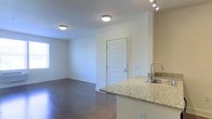 This well-appointed single-floor space offers a seamless open-concept design that connects the kitchen and living areas, creating an inviting and functional layout. The kitchen features elegant granite countertops with subtle veining, a stainless steel sink with a modern arc-style faucet, and track lighting overhead, providing both style and practicality. The countertop extends as a peninsula, subtly defining the kitchen space while maintaining an open flow into the living area.

Rich, dark hardwood flooring runs throughout the main living space, adding warmth and a polished aesthetic. The living area is generously proportioned, bathed in natural light streaming through a window along the far wall. Neutral, light-toned walls complement the flooring and contribute to the overall bright and airy ambiance of the space.

Flush-mount ceiling light fixtures provide consistent, even illumination throughout. The entryway features a clean, paneled white door with a contemporary lever-style handle, framed neatly against the neutral walls. Crisp white baseboards and trim throughout add a refined finishing touch. Overall, the space presents a modern, move-in-ready environment with a cohesive design aesthetic that balances comfort and contemporary style.