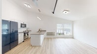 This single-floor loft-style unit blends industrial character with clean, modern design. The open-concept living and kitchen area is the centerpiece of the space, distinguished by soaring vaulted ceilings, exposed dark steel beams and piping, and light wood-look flooring that runs seamlessly throughout. Abundant natural light filters in through a large window in the living area, enhancing the bright, airy atmosphere created by the crisp white walls and high ceilings.

The kitchen is well-appointed with white cabinetry, stainless steel appliances including a range and microwave, a sleek gray countertop island with a sink, and pendant lighting overhead. A bold dark-finish refrigerator adds a striking contrast to the otherwise light palette.

The bedroom offers generous vertical space, complete with a ceiling-mounted ring pendant light, exposed piping accents, a full closet with panel doors, and a window bringing in natural light. An additional bedroom features vaulted ceilings and an outdoor-facing window with a wall-mounted HVAC unit.

The hallway connecting the rooms is accented by a decorative globe wall sconce, adding a refined industrial touch. The full bathroom features warm earth-toned tile surrounding a soaking tub and shower combination, a wood-finish vanity, white countertop, and light wood flooring.