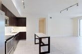 This single-floor space showcases a contemporary open-plan layout that seamlessly integrates the kitchen and living area. The kitchen is outfitted with rich, dark espresso-toned cabinetry that extends to the ceiling, providing ample storage. Light-colored countertops offer a striking contrast against the dark cabinetry, while stainless steel appliances, including a range and dishwasher, add a polished, modern touch. An undermount sink is positioned along the cabinetry run, complemented by under-cabinet lighting that brightens the workspace.

The open living area flows naturally from the kitchen, unified by large-format light-toned tile flooring that runs throughout the space, contributing to a clean and airy aesthetic. A freestanding dark-framed table with a light top serves as a functional centerpiece, echoing the kitchen's color palette. Track lighting installed on the ceiling provides adjustable illumination across both zones.

The crisp white walls and bright ceiling enhance the sense of openness, while a doorway visible in the background leads to additional areas of the unit. Overall, the space presents a cohesive, modern design with a neutral palette and clean lines.