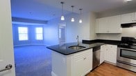 This single-floor residence offers a clean, modern layout designed for comfortable everyday living. The open-concept kitchen serves as a central hub of the home, featuring a large island with sleek dark granite countertops, a stainless steel undermount sink, and white shaker-style cabinetry. Three pendant lights hang above the island, providing focused task lighting while adding a stylish touch. A stainless steel dishwasher is seamlessly integrated into the cabinetry. The kitchen flows naturally into a spacious carpeted living area, creating an inviting, connected atmosphere well-suited for both relaxing and entertaining. A bedroom, accessible via the main living space, is finished with soft carpet flooring and neutral gray walls. Two windows fitted with white horizontal blinds allow generous natural light to pour in, offering views of the surrounding greenery and creating a bright, airy ambiance. Clean white baseboards and crisp door trim run throughout the space, lending a polished, well-maintained appearance. The overall design is understated and functional, with a neutral color palette that provides a versatile backdrop for a variety of interior styles.