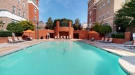 This inviting space centers around a generously sized outdoor swimming pool, framed by stately multi-story brick buildings that lend a sense of classic architectural grandeur. The pool deck is lined with lounge chairs and shaded by vibrant orange umbrellas, creating an ideal setting for relaxation. A decorative brick feature wall anchors the far end of the pool area, accented by ornamental columns and lush greenery, while classic lamp posts add a timeless charm to the surroundings. The open-air courtyard benefits from abundant natural light and open sky views.

The interior living spaces are finished with a warm, neutral palette throughout. Plush carpeting flows through the bedroom areas, which feature windows overlooking green outdoor scenery, filling the rooms with natural light. Each bedroom is accompanied by a well-appointed bathroom showcasing rich dark-stained wood cabinetry with multiple drawers, granite-style countertops, a large mirror, and a tub-shower combination set on warm hardwood flooring. The kitchen area, visible from the living space, features pendant lighting and a breakfast bar, complementing the open and functional layout. Clean white trim and understated finishes throughout give the interior a polished yet comfortable feel.