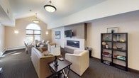 This single-floor gathering space offers a warm and inviting atmosphere designed for comfort and social interaction. The room is anchored by a sleek, tile-surround fireplace with a wall-mounted flat-screen television above, creating a natural focal point for the seating area. A generous arrangement of sofas, armchairs, and accent chairs — upholstered in neutral and patterned fabrics — provides flexible seating options throughout the space.

Vaulted ceilings lend a sense of openness and grandeur, while a striking arched window at one end floods the room with natural light, enhancing the bright, airy ambiance. Pendant and drum-shade lighting fixtures complement the natural light, ensuring the space remains well-lit and welcoming at all hours.

Dark, patterned carpeting runs throughout, grounding the room with a classic, polished aesthetic that contrasts neatly with the warm beige and taupe wall tones. A dedicated work or dining area features a compact table and chairs positioned near windows with slatted blinds, offering a functional nook within the larger space.

Wall sconces, decorative artwork, and thoughtfully placed accent lamps add character and warmth. A bookshelf with decorative items and a corkboard near the entrance contribute to the space's community-oriented character, suggesting a shared lounge setting suited for relaxation, socializing, or quiet work.