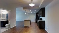 This single-floor residence showcases a thoughtfully designed open-concept layout that seamlessly connects the kitchen and living areas, creating a bright and cohesive living environment. The kitchen is a standout feature, outfitted with dark espresso cabinetry that extends to the ceiling, providing ample storage while lending a bold, contemporary aesthetic. Granite countertops in earthy tones complement the cabinetry and add a sense of sophistication. A central island serves as both a functional workspace and a natural gathering point, complete with a sink and pendant lighting that adds warmth and visual interest overhead. Stainless steel appliances, including a range and microwave, are seamlessly integrated into the cabinetry along the perimeter wall. Rich hardwood-style flooring flows throughout the space, grounding the interior with a warm, inviting tone. Recessed lighting and a decorative flush-mount fixture ensure the space is well-lit throughout the day. The open living area beyond the kitchen offers generous room to arrange furnishings freely, with soft neutral wall tones creating a clean, versatile backdrop. The overall ambiance is modern and functional, balancing stylish finishes with practical, everyday livability.