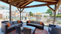 The Grill Area & Pavilion is a well-appointed outdoor amenity space designed for relaxed gatherings and al fresco entertaining. At its centerpiece stands a striking wood-beamed gazebo with an exposed timber-frame ceiling, sheltering a generous lounge area furnished with cushioned wicker sofas and armchairs accented with orange throw pillows, arranged around a low coffee table atop a patterned area rug. Open on all sides, the pavilion offers unobstructed views of the community pool, complete with lounge chairs and yellow umbrellas, creating a seamless indoor-outdoor connection. Adjacent to the pavilion, a covered patio extends the usable outdoor space, featuring high-top metal dining tables and chairs positioned alongside large windows that look into an interior common area. A ceiling fan overhead adds comfort, and a beverage vending machine provides convenient refreshments. Brick-accented columns and white-painted woodwork give the patio a classic, residential character. The surrounding pool deck is landscaped with mature trees, colorful plantings, and a pergola structure, all bathed in natural sunlight. Together, these spaces create a vibrant outdoor retreat ideal for socializing, grilling, and enjoying warm-weather amenities.