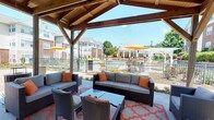 The Grill Area & Pavilion is a well-appointed outdoor amenity space designed for relaxed gatherings and al fresco entertaining. At its centerpiece stands a striking wood-beamed gazebo with an exposed timber-frame ceiling, sheltering a generous lounge area furnished with cushioned wicker sofas and armchairs accented with orange throw pillows, arranged around a low coffee table atop a patterned area rug. Open on all sides, the pavilion offers unobstructed views of the community pool, complete with lounge chairs and yellow umbrellas, creating a seamless indoor-outdoor connection. Adjacent to the pavilion, a covered patio extends the usable outdoor space, featuring high-top metal dining tables and chairs positioned alongside large windows that look into an interior common area. A ceiling fan overhead adds comfort, and a beverage vending machine provides convenient refreshments. Brick-accented columns and white-painted woodwork give the patio a classic, residential character. The surrounding pool deck is landscaped with mature trees, colorful plantings, and a pergola structure, all bathed in natural sunlight. Together, these spaces create a vibrant outdoor retreat ideal for socializing, grilling, and enjoying warm-weather amenities.