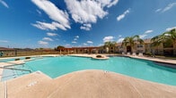 This outdoor pool area is a well-appointed recreational amenity designed with a resort-style aesthetic. The centerpiece is a generously sized freeform swimming pool with clear turquoise water, accented by blue tile detailing along the pool's edge and equipped with stainless steel entry rails for easy access. The pool features a shallow wading area and a small landscaped island focal point within the water, adding visual interest to the layout.

A striking wood-beam pergola structure anchors the poolside deck, providing shaded seating and gathering space beneath an open sky. The surrounding concrete deck offers ample room for lounge chairs and outdoor furniture, making it ideal for relaxation and socializing.

Adjacent to the pool stands a Mediterranean-inspired clubhouse building, characterized by a terracotta tile roof, arched entryways, and stucco exterior walls. Tall palm trees are strategically placed throughout the area, enhancing the tropical ambiance. The entire pool area is enclosed by a wrought iron fence, ensuring safety and privacy.

Under open skies with sweeping views and abundant natural sunlight, this outdoor space delivers a bright, welcoming atmosphere suited for year-round enjoyment in a warm climate.