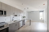 Unit 504 at 1213–1221 Broad St is a thoughtfully designed one-bedroom, one-bathroom residence that makes excellent use of its compact footprint across a single floor. Light hardwood flooring flows throughout, complementing a clean, neutral color palette that enhances the sense of openness.

The kitchen is a standout feature, outfitted with full-height wood-grain cabinetry in a warm gray-brown tone, light quartz countertops, and a white beveled subway tile backsplash. A complete suite of stainless steel appliances — including a refrigerator, electric range, over-the-range microwave, and dishwasher — adds both functionality and a polished, modern aesthetic. A matte black faucet at the undermount sink provides a striking contrast to the bright counters.

The bedroom is a well-proportioned retreat, featuring a large window that invites generous natural light and offers open views of the surrounding cityscape and treetops. Double shaker-style closet doors with matte black hardware provide ample storage, while the light hardwood floors and soft gray walls create a calm, comfortable atmosphere.

The bathroom is finished with consistent modern detailing, including matte black grab bars and fixtures that echo the hardware found throughout the unit.

White shaker-style interior doors with matte black lever hardware are used consistently throughout, tying the space together with a cohesive, contemporary design sensibility.