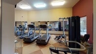 This fitness center is thoughtfully designed to support a full range of workout activities within a single-floor facility. The entrance greets visitors with soaring vaulted ceilings, warm neutral-toned walls, and large-format tile flooring, creating an open and welcoming atmosphere. A branded entry mat and glass door allow natural light to filter into the lobby area, setting an inviting tone upon arrival.

The main workout area is defined by a striking combination of warm yellow and deep red accent walls, complemented by a diamond-patterned rubber and tile floor that adds both visual interest and functional durability. Full-length mirrors line one wall, amplifying the sense of space and allowing users to monitor their form. The room is equipped with multiple treadmills and cardiovascular machines, as well as a commercial-grade Life Fitness multi-station strength machine, a dumbbell rack with a full range of free weights, and adjustable benches. Overhead fluorescent lighting ensures the space is consistently well-lit.

A corridor connecting the workout area to the exit features an arched architectural detail, adding a refined design element to the functional space. This hallway also houses a water cooler station and provides access to an ADA-accessible restroom. The overall layout flows intuitively, balancing practicality with a clean, professional aesthetic suited for daily fitness use.