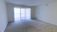This single-floor space centers around a generously sized living area finished with wall-to-wall carpeting in a neutral tone. The room is defined by its large sliding glass door, fitted with vertical blinds, which serves as the primary source of natural light and provides direct access to an outdoor balcony. The bright, white walls and textured ceiling contribute to an open, airy feel throughout the space. A standard interior door on one side of the living area leads to an adjoining room, while an additional recessed panel door is visible along another wall, likely providing access to a utility or storage area. A floor-level HVAC vent is positioned beneath the sliding door, ensuring climate control is efficiently distributed across the room. The overall design is straightforward and functional, with a clean, neutral palette that offers flexibility for a variety of furnishing arrangements. The abundance of natural light filtering through the sliding glass door enhances the sense of openness, making the living area feel bright and inviting despite its simple, understated finishes.