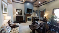 This single-story space showcases a generously proportioned living area designed with classic, traditional aesthetics. The room immediately draws attention to its dramatically tall ceilings, which are accented by coffered detailing and crown molding, adding a sense of architectural refinement. Dark hardwood floors run throughout, providing a rich contrast to the neutral, light-toned walls painted in a soft blue-gray hue.

A central fireplace serves as the focal point of the room, framed by a wood mantel and a light stone surround, with a mounted flat-screen television positioned above. Flanking the fireplace are large windows dressed with patterned blue drapes and wooden blinds, allowing for controlled natural light. Recessed lighting and table lamps contribute to a warm, layered lighting scheme throughout the space.

The furnishings are traditional in style, featuring deep brown leather seating, upholstered accent chairs with geometric patterns, and dark wood accent pieces including a sideboard with a decorative mirror. A large area rug anchors the seating arrangement, completing the cohesive, inviting ambiance of the room.