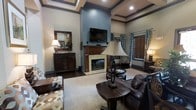 This single-story space showcases a generously proportioned living area designed with classic, traditional aesthetics. The room immediately draws attention to its dramatically tall ceilings, which are accented by coffered detailing and crown molding, adding a sense of architectural refinement. Dark hardwood floors run throughout, providing a rich contrast to the neutral, light-toned walls painted in a soft blue-gray hue.

A central fireplace serves as the focal point of the room, framed by a wood mantel and a light stone surround, with a mounted flat-screen television positioned above. Flanking the fireplace are large windows dressed with patterned blue drapes and wooden blinds, allowing for controlled natural light. Recessed lighting and table lamps contribute to a warm, layered lighting scheme throughout the space.

The furnishings are traditional in style, featuring deep brown leather seating, upholstered accent chairs with geometric patterns, and dark wood accent pieces including a sideboard with a decorative mirror. A large area rug anchors the seating arrangement, completing the cohesive, inviting ambiance of the room.
