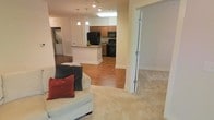 This single-floor residence showcases an open-concept layout that connects the living area to the kitchen, creating a cohesive and functional space. The living area features plush, light-colored carpeting that adds warmth underfoot, while the kitchen and adjoining areas transition to hardwood-style flooring, providing a clean visual distinction between spaces. Pendant lighting hangs above a kitchen island with a stone-look countertop, serving as both a functional workspace and a natural divider between the cooking and living zones. The kitchen is equipped with medium-toned wooden cabinetry, black appliances including a refrigerator, stove, and built-in microwave, offering a classic and practical cooking environment. Recessed lighting throughout the ceiling ensures consistent illumination across the open floor plan. A doorway visible to the right, fitted with a glass-paneled door, leads to an additional room, likely a bedroom, featuring matching light carpeting. The overall ambiance is warm and inviting, with neutral tones throughout the walls and flooring creating a versatile backdrop suitable for a variety of interior styles.