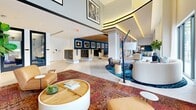 Located at 501 NW Seventh Ave in Fort Lauderdale, FL, this expansive single-floor amenity and leasing space offers a thoughtfully curated environment that blends functionality with artistic flair. The layout spans a generous footprint and includes 14 distinct areas, among them two dedicated private offices and a variety of multi-use common spaces.

The main lobby commands immediate attention with soaring double-height ceilings, floor-to-ceiling windows dressed in sheer white drapery, and an abundance of natural light that floods the interior. A long, light wood workstation anchors the leasing area, outfitted with computer workstations, pendant globe lighting, and warm cognac leather seating. Vibrant, patchwork-style area rugs in rich jewel tones define each zone, contrasting beautifully with large-format white tile flooring.

A lounge area features a curved cream sectional sofa, eclectic accent chairs, and a bold teal abstract area rug, with sliding glass doors opening to an outdoor terrace. A gallery corridor showcases framed black-and-white photography mounted on deep teal accent walls, alongside a large-scale colorful mural that energizes the space. A wood-paneled ceiling section adds warmth to the mailroom area. Private glass-walled offices offer an intimate, contemporary work setting. Throughout, lush indoor plants and curated artwork reinforce an inviting, design-forward atmosphere.