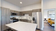 This contemporary single-floor residence showcases a clean, modern design throughout, with crisp white walls, wood-tone flooring, and an abundance of natural light. The kitchen is a standout space, featuring a generous waterfall-edge quartz island, wood-grain cabinetry extending to the ceiling, and a full suite of stainless steel appliances including a gas range, built-in microwave, dishwasher, and French door refrigerator. The living area is bright and welcoming, anchored by large black-framed glass doors that open directly to the outdoors, framing views of mature trees and greenery. A dedicated office or flex room continues the nature-connected theme with an oversized window offering tranquil outdoor views, making it an ideal workspace. The bedrooms are serene and light-filled, each featuring sizable windows with tree-line views and warm hardwood-style flooring. The bathrooms are finished with wood-grain vanities, white quartz countertops, and clean tile work — one featuring a full soaking tub and shower combination, and another with direct access to a walk-in closet with built-in shelving. Throughout, the space maintains a cohesive aesthetic of modern minimalism, blending functionality with refined residential comfort.
