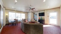 This single-floor community space is designed to offer a comfortable and sociable environment for its occupants. The main lounge area is anchored by a generously sized curved sectional sofa in a warm taupe tone, providing ample seating and encouraging conversation. Recessed lighting and a ceiling fan overhead ensure the space remains bright and well-ventilated throughout the day.

A wall-mounted flat-screen television is positioned opposite the seating area, accompanied by a dark wood media console adorned with colorful decorative vases in shades of amber, teal, and green. Vibrant vertical artwork panels featuring bold splashes of color add an artistic focal point to the room, complementing the neutral cream-toned walls and deep red carpeting.

Large windows along one wall invite an abundance of natural light, offering views of the outdoor surroundings and creating an open, airy atmosphere. A secondary seating area near the windows features wicker-style chairs and a dining table, making it ideal for both casual gatherings and quiet relaxation. Architectural details such as crown molding and column accents lend a refined, polished character to the overall design. The space strikes a thoughtful balance between comfort and style, making it a functional and welcoming common area.