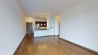 This single-floor space showcases an open-concept layout that seamlessly connects the living area to the kitchen, creating a sense of spaciousness and flow. The warm-toned hardwood flooring extends throughout the main living area, adding both visual warmth and a polished aesthetic to the interior.

The kitchen is a standout feature, equipped with dark wood cabinetry that contrasts elegantly against the white peninsula counter, which offers additional workspace and casual seating potential. Pendant lighting fixtures hang above the peninsula, providing both task lighting and a decorative focal point. A stainless steel refrigerator is visible, complementing the modern design direction of the kitchen. Track lighting above the kitchen further enhances the illumination of the cooking and prep area.

The living area is generously proportioned, offering flexible possibilities for furniture arrangement. White walls and a neutral ceiling keep the space feeling bright and airy. A paneled entry door is visible toward the back, providing a defined point of entry. A hallway leads to additional areas of the unit, suggesting further rooms beyond the main living space. Overall, the space presents a clean, contemporary aesthetic with functional design elements throughout.