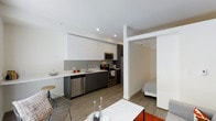This single-floor unit offers a thoughtfully designed open-concept layout that seamlessly integrates the kitchen, dining, and living areas into one cohesive space. The modern kitchen is a standout feature, outfitted with two-tone cabinetry — crisp white upper cabinets contrasting with dark lower cabinets — paired with stainless steel appliances including a dishwasher, range, and over-the-range microwave. A clean, light-toned countertop runs along the kitchen, complemented by a simple yet functional sink setup and a subtle gray tile backsplash. The flooring throughout is a warm, wood-look material that adds texture and continuity across the open living space. Recessed lighting in the ceiling provides bright, even illumination throughout. The living area flows naturally from the kitchen, offering space for dining and relaxation. A sliding door or open passage leads to a separate bedroom, visible in the background, providing privacy while maintaining the unit's open feel. The overall aesthetic is clean, contemporary, and functional, with a neutral color palette accented by modern furnishings and décor touches that enhance the minimalist design sensibility.