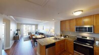 This thoughtfully designed single-floor residence offers a seamless open-concept layout that connects the kitchen, dining area, and living room into one cohesive, light-filled space. The kitchen is outfitted with warm honey-toned cabinetry, granite countertops, a stainless steel sink set into a peninsula, and a full suite of stainless steel appliances including a range, microwave, dishwasher, and refrigerator with ice maker. The peninsula provides an ideal transition into the dining area, where a round table with seating for four sits adjacent to the living room. The living area is anchored by rich hardwood flooring, crown molding, and large sliding glass doors that open to a private balcony with outdoor views, flooding the space with natural light. The bedroom is generously proportioned, featuring plush carpeting, crown molding, and its own sliding glass door access to the outdoors. The full bathroom is finished with warm neutral tones, a wood-front vanity with granite countertop, a large framed mirror, and a shower-tub combination. Throughout the residence, neutral wall tones, recessed and flush-mount lighting, and consistent warm wood accents create a cohesive and inviting atmosphere.