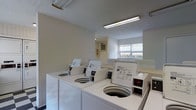 This single-floor laundry facility is designed to serve multiple users efficiently. The room features a row of top-loading washing machines lined along the walls, providing ample washing capacity. The space is illuminated by fluorescent overhead lighting, ensuring bright and functional visibility throughout. A window along one wall allows natural light to filter in, offering a view of outdoor greenery and contributing to a more open feel. The walls are finished in a clean, neutral tone, and the overall layout is straightforward and practical. Additional equipment, including what appears to be a dryer unit, is positioned nearby, rounding out the laundry amenities. A wall-mounted utility panel is also visible, suggesting the room is well-equipped for its purpose. The space is clean and organized, making it a convenient and accessible shared facility.