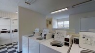 This single-floor laundry facility is designed to serve multiple users efficiently. The room features a row of top-loading washing machines lined along the walls, providing ample washing capacity. The space is illuminated by fluorescent overhead lighting, ensuring bright and functional visibility throughout. A window along one wall allows natural light to filter in, offering a view of outdoor greenery and contributing to a more open feel. The walls are finished in a clean, neutral tone, and the overall layout is straightforward and practical. Additional equipment, including what appears to be a dryer unit, is positioned nearby, rounding out the laundry amenities. A wall-mounted utility panel is also visible, suggesting the room is well-equipped for its purpose. The space is clean and organized, making it a convenient and accessible shared facility.