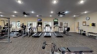 Located at 3217 Ramsgate Rd in Augusta, Georgia, this fitness facility offers a comprehensive and thoughtfully organized workout environment across a single floor. The space is finished with warm yellow walls, crown molding, and a textured ceiling fitted with recessed lighting and ceiling fans, creating a comfortable and well-ventilated atmosphere throughout.

The cardio zone features multiple treadmills, an elliptical, and a recumbent bike, each equipped with individual mounted flat-screen monitors. Large glass French doors along this wall flood the area with natural light and provide views of an outdoor patio area. Ceiling fans overhead ensure air circulation during intense workouts.

The strength training section is generously stocked with a cable machine, lat pulldown station, functional trainer, and a power rack, offering a full range of resistance training options. A dedicated free weights area includes a comprehensive dumbbell rack and a flat bench, complemented by a large wall mirror that enhances the sense of space and allows for form checks.

A functional training corner rounds out the facility with kettlebells, medicine balls, plyo boxes, resistance bands, stability balls, and foam rollers. A wall-mounted TV and motivational wall decal add personality to this zone. A small workstation desk area is also present, suggesting space for personal training consultations or administrative use.