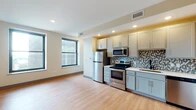 This single-floor unit offers a clean, modern aesthetic defined by light hardwood flooring and neutral warm-toned walls that flow consistently throughout the space. The open-concept kitchen and living area serves as the heart of the unit, where large black-framed windows invite abundant natural light and offer views of the surrounding urban streetscape. The kitchen is outfitted with gray shaker-style cabinetry, a full suite of stainless steel appliances including a refrigerator, electric range, built-in microwave, and dishwasher, as well as light-toned countertops and a geometric mosaic tile backsplash that adds a contemporary decorative accent. Recessed lighting illuminates the kitchen and living zones, enhancing the open and airy feel of the combined space. Additional rooms feature the same hardwood flooring and black-framed windows, maintaining a cohesive look throughout. The bathroom is thoughtfully finished with a rich wood-toned vanity, white quartz countertop, an undermount sink with oil-rubbed bronze fixtures, a backlit LED vanity mirror, and a tiled tub-shower combination. The overall design balances functionality with modern style, resulting in a bright, move-in ready space.