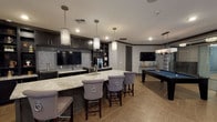 The Gentry's single-floor amenity lounge is a thoughtfully designed social space that seamlessly integrates multiple entertainment and relaxation zones. The layout flows naturally from a well-appointed wet bar area through a game room and into a comfortable lounge, creating distinct yet connected environments for residents to enjoy.

The wet bar serves as a central hub, anchored by a generously sized granite-topped island with an undermount sink and seating for multiple bar-height chairs upholstered in gray velvet with nailhead trim and ring pulls. Dark espresso cabinetry lines the walls, complemented by a shimmering mosaic tile backsplash and a wall-mounted television. Open shelving provides display space, and a beverage station adds convenience. Cylindrical pendant lights illuminate the island, contributing to the upscale atmosphere.

Adjacent to the bar, a billiards table with a blue felt surface offers recreational entertainment, accompanied by additional wall-mounted televisions. The dining area features high-top tables beside floor-to-ceiling windows framed by bold patterned drapery, flooding the space with natural light and offering views of lush outdoor greenery.

The lounge area centers around a marble-surround fireplace flanked by built-in dark shelving, creating a sophisticated focal point. Tufted seating, a chevron-patterned area rug, and curated décor complete the inviting atmosphere. Throughout, herringbone-laid wood-look flooring unifies the entire space.