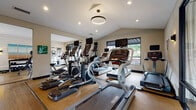 Located at 300 Royal Oaks Blvd in Franklin, Tennessee, this single-floor fitness center is thoughtfully designed to support a well-rounded workout experience. The space is organized into distinct zones, allowing for a seamless flow between cardio and strength training areas. The main cardio zone features a row of treadmills and elliptical trainers, each equipped with individual display screens, positioned alongside windows that bring in natural light and views of the surrounding greenery. Overhead, a circular pendant light fixture complements the recessed lighting throughout, creating a bright and energizing atmosphere. The flooring transitions between hardwood-style planks and rubberized mat surfaces, providing both durability and comfort underfoot. A dedicated strength training area houses a dumbbell rack with a full range of free weights, a weight bench, and cable/resistance machines, all set against a shiplap-accented wall adorned with colorful artwork. A water rowing machine is positioned near the entrance, adding a low-impact full-body training option. Large steel-framed glass partitions offer a transparent connection to an adjacent lounge or common area, enhancing the open feel of the space. Wall-mounted sconce lighting and subtle decor contribute to the polished, modern aesthetic throughout.