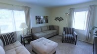 This single-story space presents a thoughtfully arranged living area designed for comfort and functionality. The room features neutral wall tones complemented by plush, wall-to-wall carpeting that adds warmth underfoot. Natural light filters in generously through two windows dressed with elegant floor-length curtains in a soft cream hue, creating a bright and inviting atmosphere.

The seating arrangement is both practical and stylish, centered around a spacious gray upholstered sofa accented with patterned throw pillows. A matching tufted ottoman serves as a versatile centerpiece, doubling as a coffee table or additional seating. A bold patterned accent chair in dark tones provides a visual contrast and adds character to the room.

Decorative elements include an abstract canvas print on the wall and a sculptural metallic wall accent, lending the space a contemporary aesthetic. A sleek floor lamp in the corner provides additional ambient lighting, while a side table with a table lamp and small shelving below offers a convenient surface for everyday items. Overall, the living area balances casual comfort with understated modern style.