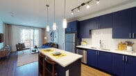 This thoughtfully designed single-floor residence seamlessly blends contemporary style with functional living. The open-concept kitchen is a standout feature, showcasing bold navy blue cabinetry that extends from floor to ceiling, complemented by white subway tile backsplash, sleek white quartz countertops, and a full suite of stainless steel appliances including a refrigerator, dishwasher, microwave, and range. A central kitchen island with bar seating and pendant lighting overhead creates an inviting space for casual dining and entertaining. Warm hardwood-style flooring flows throughout the kitchen and into the connected living area, which is bathed in natural light from large sliding glass doors that open to an outdoor balcony with views of the surrounding community. The living room offers a comfortable, open layout ideal for relaxing or hosting guests. The bedrooms are generously sized, each featuring plush carpeting, ceiling fans, large windows with exterior views, and ample natural light. The bathroom maintains a clean, modern aesthetic with white tile flooring, a soaking tub and shower combination, and a navy vanity with a white countertop that ties back to the kitchen's cohesive design palette. Overall, the space delivers a polished, contemporary ambiance throughout.