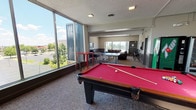Located at 111 Grafton Ave in Dayton, Ohio, this single-floor community amenity space is thoughtfully designed to offer recreation, relaxation, and fitness in one open layout. Floor-to-ceiling windows along one side of the room flood the space with natural light and provide expansive views of the surrounding Dayton skyline and tree-lined surroundings, creating an inviting, airy atmosphere throughout.

The game area serves as a central focal point, anchored by a full-size pool table with a bold red felt surface, complemented by a ping-pong table nearby — ideal for casual social gatherings. High-top bar seating with red metal stools is positioned along the window wall, offering a spot to relax while taking in the outdoor views.

A dedicated lounge area features rich brown leather sofas and armchairs arranged around a coffee table, providing a comfortable space for conversation or unwinding. The neutral carpeted flooring and two-tone gray wall treatment lend the space a modern yet welcoming feel.

A fitness zone, partially enclosed with glass partitioning, houses cardio and strength equipment including resistance training accessories, keeping active residents well-accommodated. Vending machines are conveniently available near the elevator lobby, which is accented with a distinctive herringbone-patterned feature wall, adding a refined architectural touch to the space.
