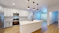 This well-appointed single-story home offers three bedrooms and two full bathrooms across a thoughtfully designed floor plan. The open-concept kitchen and living area serves as the heart of the home, centered around a large white waterfall-edge island with an undermount sink and matte black faucet. Three stylish pendant lights with brass accents hang above the island, adding a contemporary touch. The kitchen features white shaker cabinetry, a geometric hexagonal tile backsplash, stainless steel appliances including a gas range, microwave, and refrigerator, and warm wood-look flooring that extends throughout the entire home. The adjacent living area benefits from generous natural light through large windows and is equipped with a ceiling fan. The primary bedroom is a comfortable retreat with large windows, a ceiling fan, and access to a dedicated closet. The two bathrooms are finished with large-format gray tile, white vanities with LED-lit mirrors, and matte black hardware. One bathroom includes a sleek frameless sliding glass shower, while the second offers a full soaking tub. The secondary bedrooms are bright and versatile, each featuring ceiling fans and natural light. Clean white walls, modern doors with black hardware, and consistent flooring create a cohesive, contemporary aesthetic throughout.