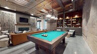 Located at 96 E Catawba St in Belmont, NC, this single-floor community space masterfully blends industrial character with refined interior design. The open layout is anchored by soaring ceilings that showcase an artistic lattice-style wood and metal ceiling treatment, exposed ductwork, and statement pendant lighting that adds warmth and texture throughout.

The centerpiece of the main gathering area is a full-size billiards table positioned beneath industrial-style hanging lights, flanked by built-in wood shelving and comfortable lounge seating. Adjacent to the game area, a cozy lounge nook features a fireplace with a reclaimed wood mantel, a wall-mounted television, and plush seating arrangements set atop layered area rugs — an inviting spot for relaxed conversation.

The dining area impresses with a stunning live-edge wood communal table surrounded by bar stools, illuminated by a sculptural brass globe chandelier. Arched decorative wall panels and built-in booth seating complete this convivial setting.

The kitchen area is defined by rich warm-toned cabinetry, white countertops, open floating shelves, and an exposed brick accent wall that lends authentic historic character. A range hood, sink, and beverage station round out the functional amenities. Polished concrete flooring flows throughout, tying each zone together in an effortlessly cohesive, contemporary-industrial aesthetic.