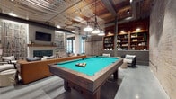 Located at 96 E Catawba St in Belmont, NC, this single-floor community space masterfully blends industrial character with refined interior design. The open layout is anchored by soaring ceilings that showcase an artistic lattice-style wood and metal ceiling treatment, exposed ductwork, and statement pendant lighting that adds warmth and texture throughout.

The centerpiece of the main gathering area is a full-size billiards table positioned beneath industrial-style hanging lights, flanked by built-in wood shelving and comfortable lounge seating. Adjacent to the game area, a cozy lounge nook features a fireplace with a reclaimed wood mantel, a wall-mounted television, and plush seating arrangements set atop layered area rugs — an inviting spot for relaxed conversation.

The dining area impresses with a stunning live-edge wood communal table surrounded by bar stools, illuminated by a sculptural brass globe chandelier. Arched decorative wall panels and built-in booth seating complete this convivial setting.

The kitchen area is defined by rich warm-toned cabinetry, white countertops, open floating shelves, and an exposed brick accent wall that lends authentic historic character. A range hood, sink, and beverage station round out the functional amenities. Polished concrete flooring flows throughout, tying each zone together in an effortlessly cohesive, contemporary-industrial aesthetic.