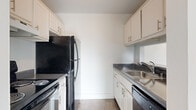 This two-floor residential space offers a well-organized and functional layout with a clean, neutral aesthetic throughout. The open-concept living and dining area is bright and airy, featuring light wood-style flooring, a ceiling fan, and large sliding glass doors dressed with vertical blinds that flood the room with natural light and provide access to outdoor greenery. A pass-through window connects the living area to the galley-style kitchen, which is efficiently designed with white cabinetry, dark granite-style countertops, and black appliances including a refrigerator, electric range, and dishwasher, all complemented by a stainless steel sink. The bedrooms are carpeted for comfort and include windows with views of surrounding greenery, offering a quiet and restful atmosphere. The bathroom features a full soaking tub with white tile surround, maintaining a clean and classic look. A dedicated in-unit laundry room houses a full-size washer and dryer with overhead shelving for added storage. An exterior staircase connects the two levels, with lush green landscaping visible from multiple vantage points, enhancing the overall sense of a connected indoor-outdoor environment.