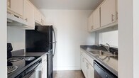 This two-floor residential space offers a well-organized and functional layout with a clean, neutral aesthetic throughout. The open-concept living and dining area is bright and airy, featuring light wood-style flooring, a ceiling fan, and large sliding glass doors dressed with vertical blinds that flood the room with natural light and provide access to outdoor greenery. A pass-through window connects the living area to the galley-style kitchen, which is efficiently designed with white cabinetry, dark granite-style countertops, and black appliances including a refrigerator, electric range, and dishwasher, all complemented by a stainless steel sink. The bedrooms are carpeted for comfort and include windows with views of surrounding greenery, offering a quiet and restful atmosphere. The bathroom features a full soaking tub with white tile surround, maintaining a clean and classic look. A dedicated in-unit laundry room houses a full-size washer and dryer with overhead shelving for added storage. An exterior staircase connects the two levels, with lush green landscaping visible from multiple vantage points, enhancing the overall sense of a connected indoor-outdoor environment.