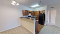 This single-floor, 2-bedroom, 2-bathroom residence features a practical and well-organized layout designed for comfortable everyday living. The kitchen is equipped with warm honey-toned wood cabinetry, dark laminate countertops, and a full suite of black appliances, including a refrigerator, range, and dishwasher. A breakfast bar peninsula separates the kitchen from the adjoining living area, which is finished with plush neutral carpeting and accented by a chandelier. The living space extends to a set of French doors with transom windows above, filling the room with natural light and providing outdoor access. Both bedrooms are generously sized, carpeted, and feature tray ceilings with large windows fitted with blinds for privacy and light control. The primary bedroom includes a ceiling fan and is connected to a bathroom with a tub-shower combination, wood vanity cabinetry, and tile flooring. A walk-in closet is accessible directly from the primary bath. The second bathroom mirrors this design with matching finishes. Throughout, the residence maintains a clean, neutral color palette with white trim, creating a bright and cohesive ambiance across all rooms.