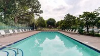 Located at 1101–1109 Northwood Dr in Eagan, Minnesota, this outdoor pool facility offers a well-appointed recreational space designed for relaxation and leisure. The centerpiece is a large, rectangular swimming pool filled with clear turquoise water, accessible via stainless steel entry rails and clearly marked with depth indicators along the pool edge.

Surrounding the pool is a spacious concrete and paved sun deck generously lined with white sling-style lounge chairs and occasional side tables, providing ample space for sunbathing and outdoor enjoyment. Patio umbrella stands are distributed throughout the deck, offering shaded seating options.

The pool area is enclosed by a sturdy green metal fence with a secured gate, ensuring safety and controlled access. Adjacent to the pool sits a single-story pool house structure with a pitched shingle roof and tan exterior siding, likely providing restroom or storage facilities for guests.

The entire setting is framed by a mature canopy of large deciduous trees, creating a natural, park-like atmosphere that offers shade and privacy. Landscaped planting beds with ornamental grasses and shrubs enhance the perimeter. The surrounding greenery and open sky contribute to a serene outdoor ambiance, making this pool area a welcoming retreat within the Eagan, MN community.