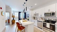 Located at 19 Harnett St in Wilmington, NC, this thoughtfully designed one-bedroom unit offers a bright, modern living experience across a single floor. The open-concept kitchen and living area serves as the heart of the space, anchored by a sleek white kitchen featuring shaker-style cabinetry with matte black hardware, a subway tile backsplash, stainless steel appliances, and a generous central island with quartz countertops and seating for two. A trio of modern pendant lights adds a striking focal point overhead. Light wood-style flooring flows throughout the kitchen and main area, enhancing the airy, contemporary feel. The living room is bathed in natural light from large windows and a glass balcony door, which opens to a private outdoor balcony with urban views. The bedroom offers a comfortable retreat with plush carpeting, a ceiling fan, and ample natural light from a large window with sheer curtains. The layout is efficient yet open, with interior doors providing access to closet and utility spaces. Clean white walls throughout the unit amplify brightness and create a neutral, versatile backdrop. The overall ambiance is modern, fresh, and welcoming — well-suited for urban living in downtown Wilmington.