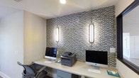 This business center offers a well-appointed, single-floor workspace designed to support productivity and professional needs. The centerpiece of the room is a long, clean-lined white workstation desk that accommodates multiple users simultaneously, outfitted with Apple iMac desktop computers and a shared multifunction printer, making it ideal for everyday business tasks. The standout design feature is the full accent wall clad in a sophisticated mosaic tile in layered shades of gray, silver, and white, lending the space a polished, contemporary aesthetic. Flanking the accent wall are two elegantly framed vertical sconce lights that cast a warm, focused glow, enhancing both the ambiance and the functionality of the workspace. The ceiling is finished in a smooth, bright white, contributing to a clean and open feel. Ergonomic task chairs provide comfortable seating at the workstation. The overall atmosphere strikes a balance between professional utility and refined design, making the business center a welcoming and efficient environment for residents or guests who need access to computing and printing resources.