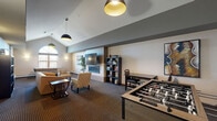 Located at 1290 County Road F W in Arden Hills, Minnesota, this single-floor community living room is a thoughtfully designed gathering space that balances comfort and entertainment. The room's most striking architectural feature is its soaring vaulted ceiling, which rises dramatically above the space and is accentuated by a large arched window that floods the area with natural light. Stylish pendant light fixtures with bold black-and-gold shades complement the warm wall sconces positioned throughout, creating a layered and inviting ambiance.

The living area is anchored by a sleek linear fireplace set against a tiled accent wall, with a wall-mounted flat-screen television above, making it an ideal spot for relaxation. Comfortable seating — including a leather sofa, patterned armchairs, and side tables — is arranged to encourage conversation and leisure. A dedicated bookshelf stocked with reading material adds a quiet, literary corner to the space.

For recreation, the room features both a foosball table and an air hockey table, along with a game and activity shelf, ensuring ample entertainment options. Patterned carpeting in deep, rich tones grounds the room, while decorative artwork and indoor greenery add warmth and personality. This versatile space is well-suited for community gatherings, casual lounging, or friendly competition.