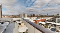 Located atop a building in Richmond, Virginia, this Sky Deck space offers a compelling blend of sophisticated interior design and an expansive open-air rooftop terrace. The interior lounge area features warm light oak hardwood flooring, deep navy-toned walls with wainscoting, and striking brushed-gold pendant lighting that lends an upscale, curated ambiance. A tufted charcoal leather sofa anchors the seating area, complemented by a bold statement floor lamp in gold. Floor-to-ceiling glass doors and windows flood the interior with natural light and frame sweeping views of the Richmond skyline, creating a seamless visual connection between inside and out. A kitchenette area is outfitted with navy cabinetry, a gold faucet, and a dramatic metallic-tiled accent wall, adding texture and a modern edge to the space. The outdoor terrace, accessed through the labeled "Sky Deck" glass entry door, is finished with herringbone-patterned pavers and furnished with bar-height tables and chairs, as well as lounge seating — all oriented to maximize the panoramic views of Richmond's downtown skyline and surrounding rooftops. The combination of refined interior finishes and an inviting outdoor terrace makes this Sky Deck an exceptional urban retreat.